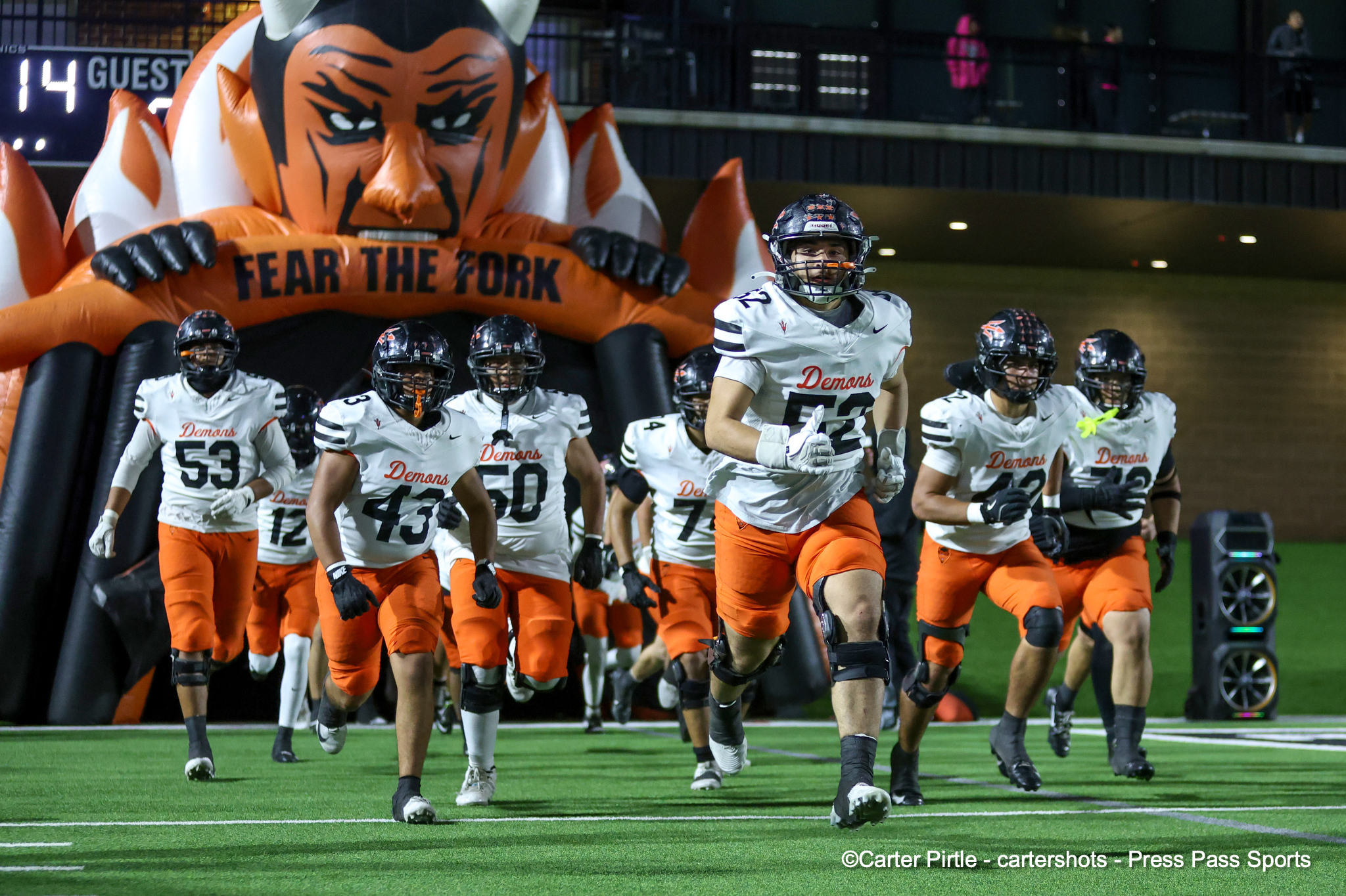 Football players in white jerseys and orange pants sprint onto the field through a large inflatable orange tiger arch reading'FEAR THE FORK'.