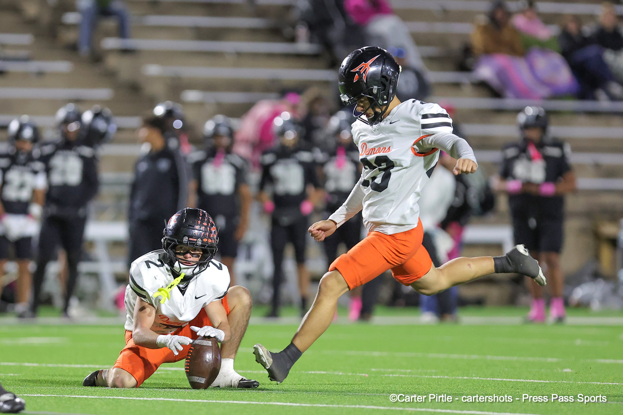 Two football players in white jerseys and orange shorts chase a fumbled ball on a bright green field as a jump occurs in the background.