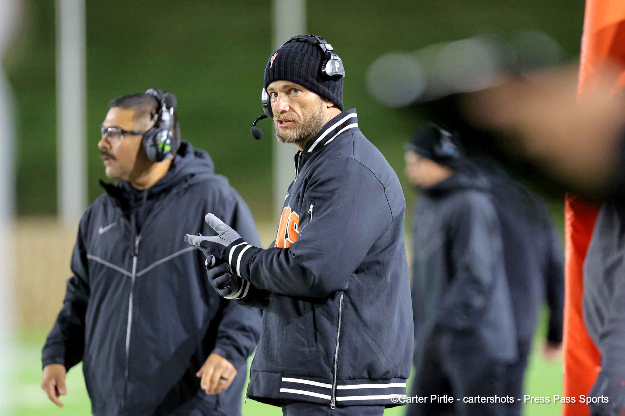 Coach wearing a black beanie and headset on the sideline during a football game, speaking to players