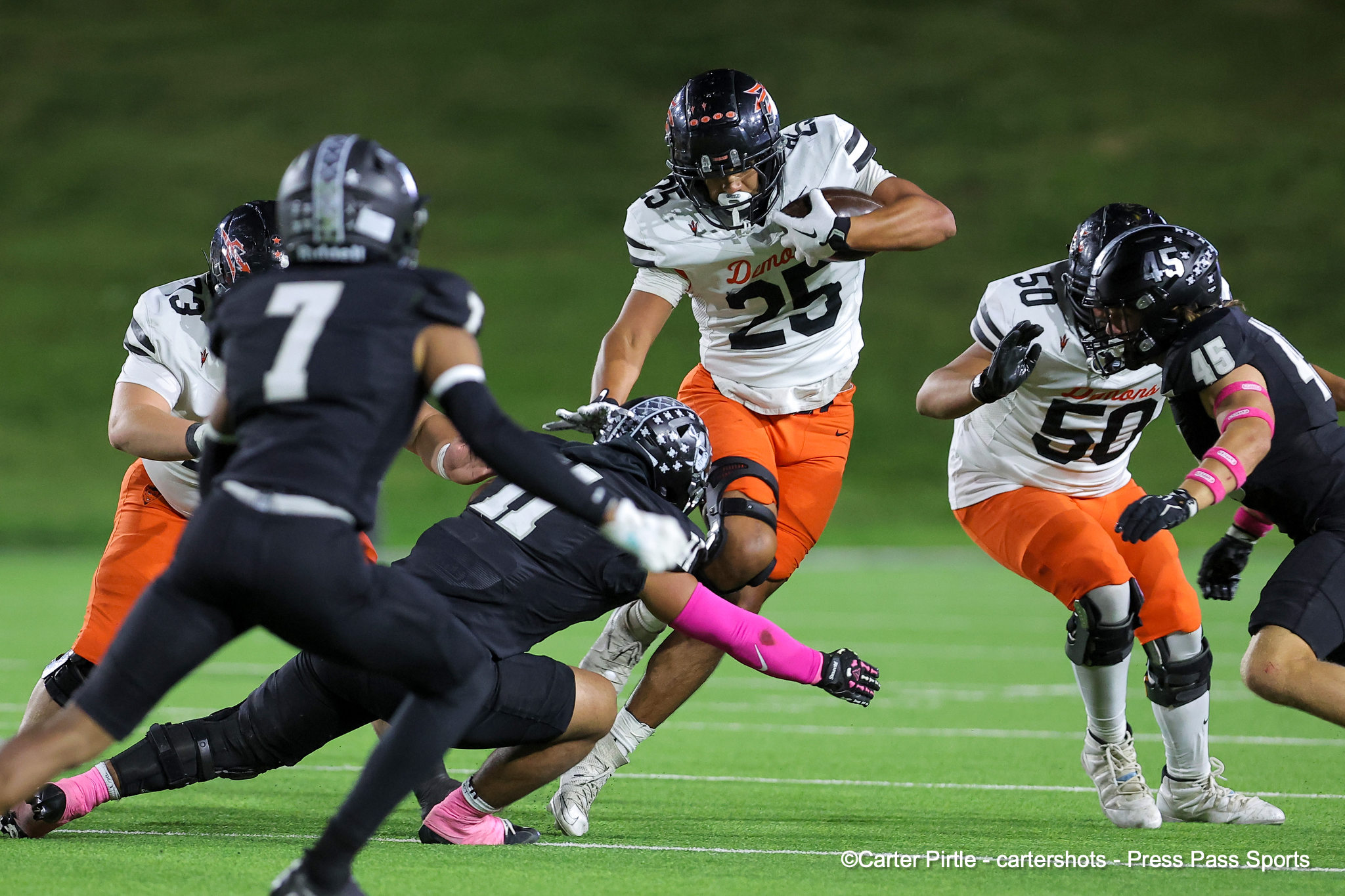 Football action: a player in white/orange holding the ball is tackled by multiple opponents on a green field at night