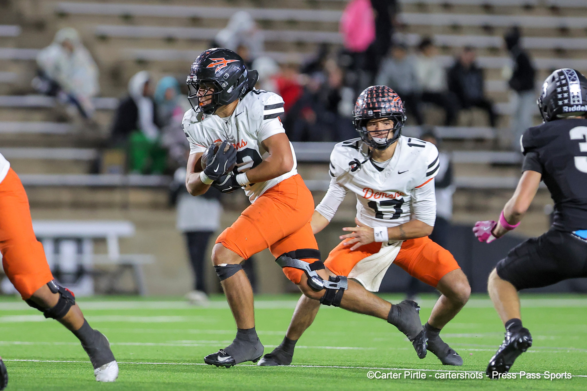 Football play: running back in white jersey and orange pants carries the ball, pursued by defenders on a green turf field.
