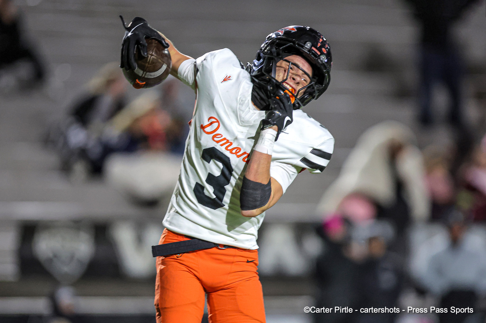 Football player in a white jersey and orange pants reaches to catch a football during a game, with a blurred crowd in the background.