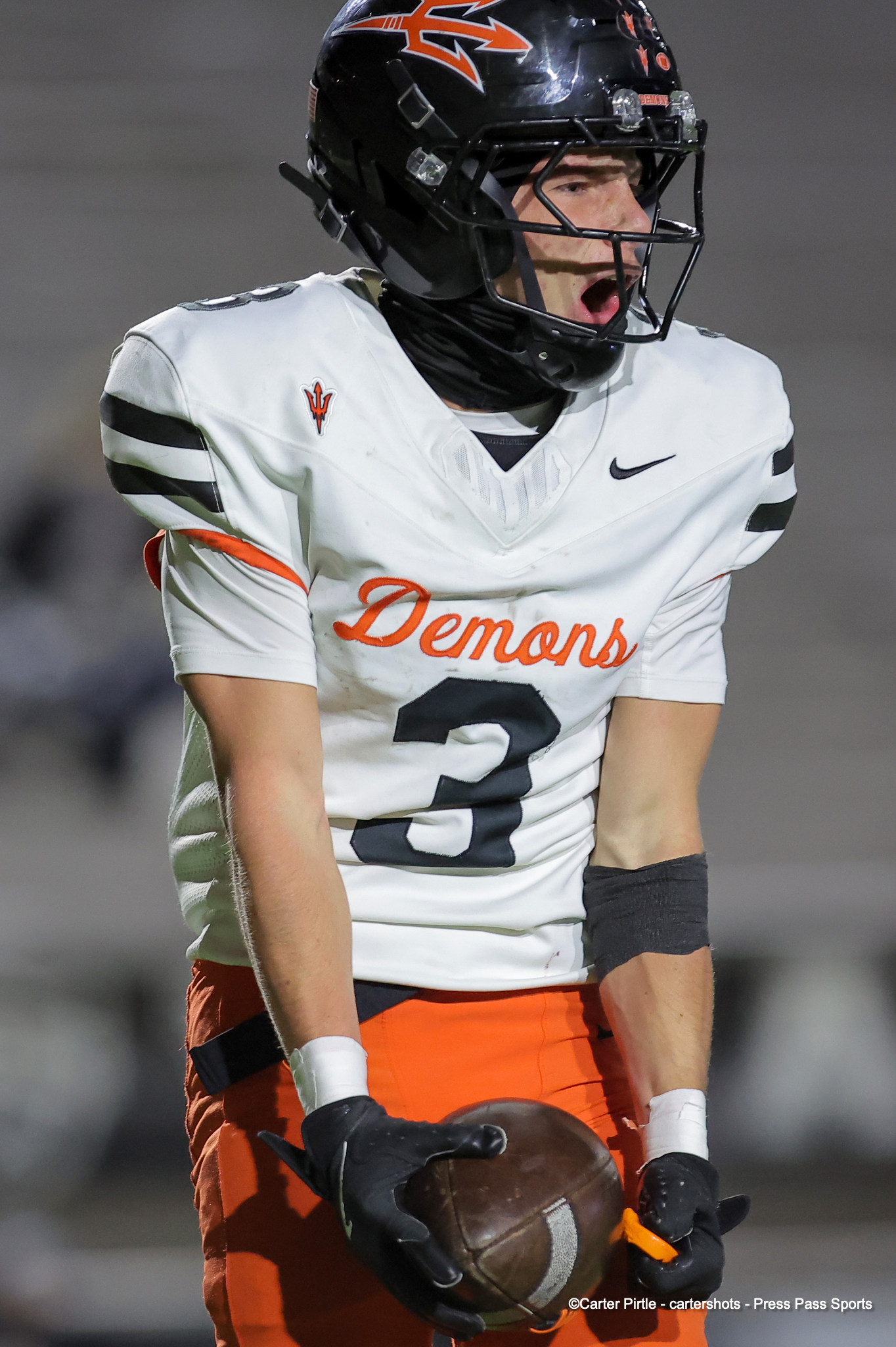 Football player in a white Demons jersey and black helmet grips a brown football during a game moment.