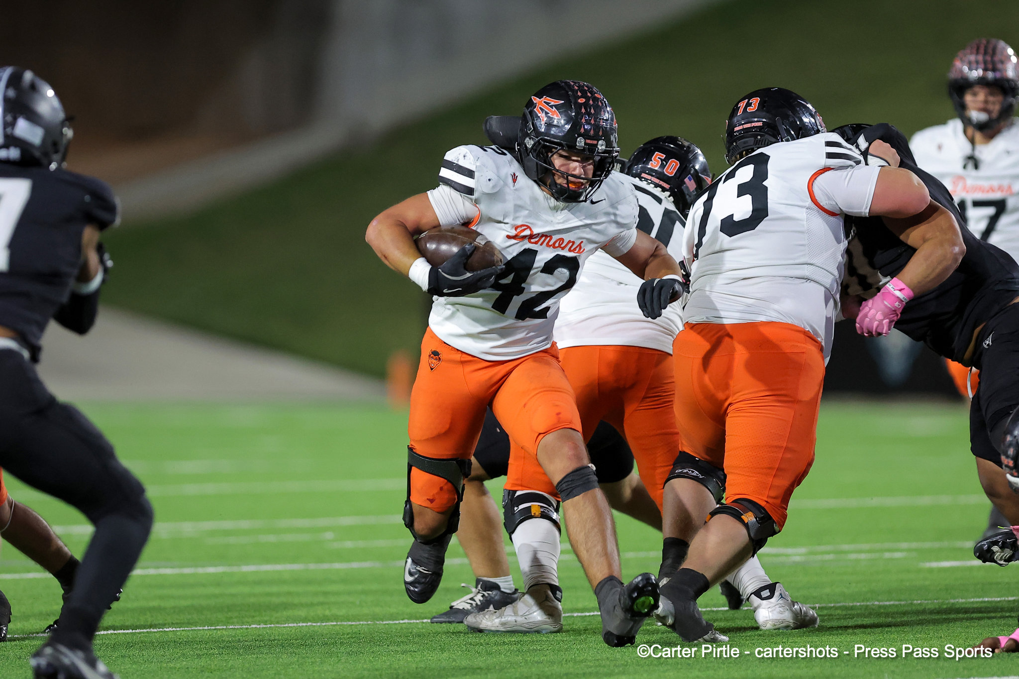 Football players in white and orange uniforms collide as a ball carrier runs with the ball on a green field at night.