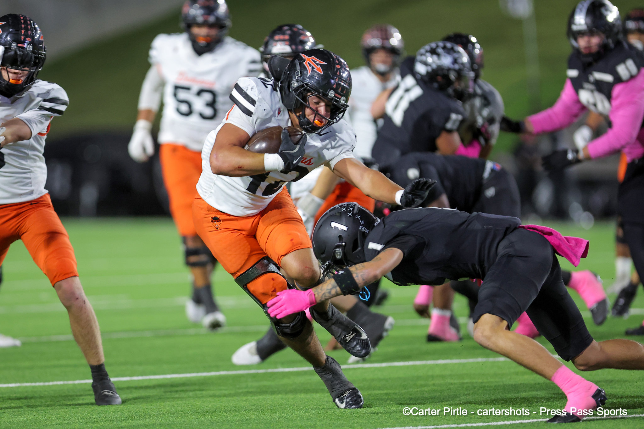 Football player in white and orange advances with the ball while pursued by opponents on a green field at night.