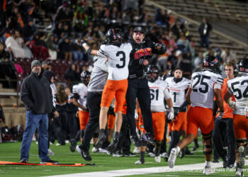 20261015-Dumas-Randall-FB-036 | Press Pass Sports Two football players in midair celebrating a win, one in black with Demons text, the other in white and orange, as teammates and a crowd cheer nearby on the field.