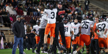 Two football players in midair celebrating a win, one in black with Demons text, the other in white and orange, as teammates and a crowd cheer nearby on the field.