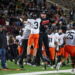 20261015-Dumas-Randall-FB-036 | Press Pass Sports Two football players in midair celebrating a win, one in black with Demons text, the other in white and orange, as teammates and a crowd cheer nearby on the field.