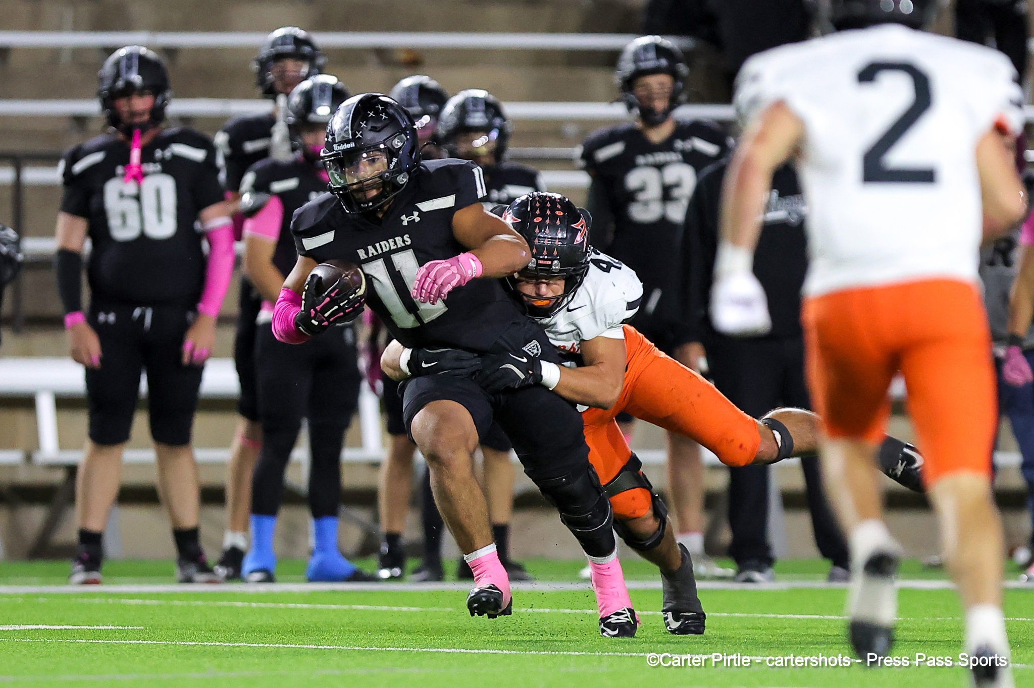 Black Raiders ball carrier dodges a tackle from a white-and-orange defender on a green football field, pink gloves visible in action.
