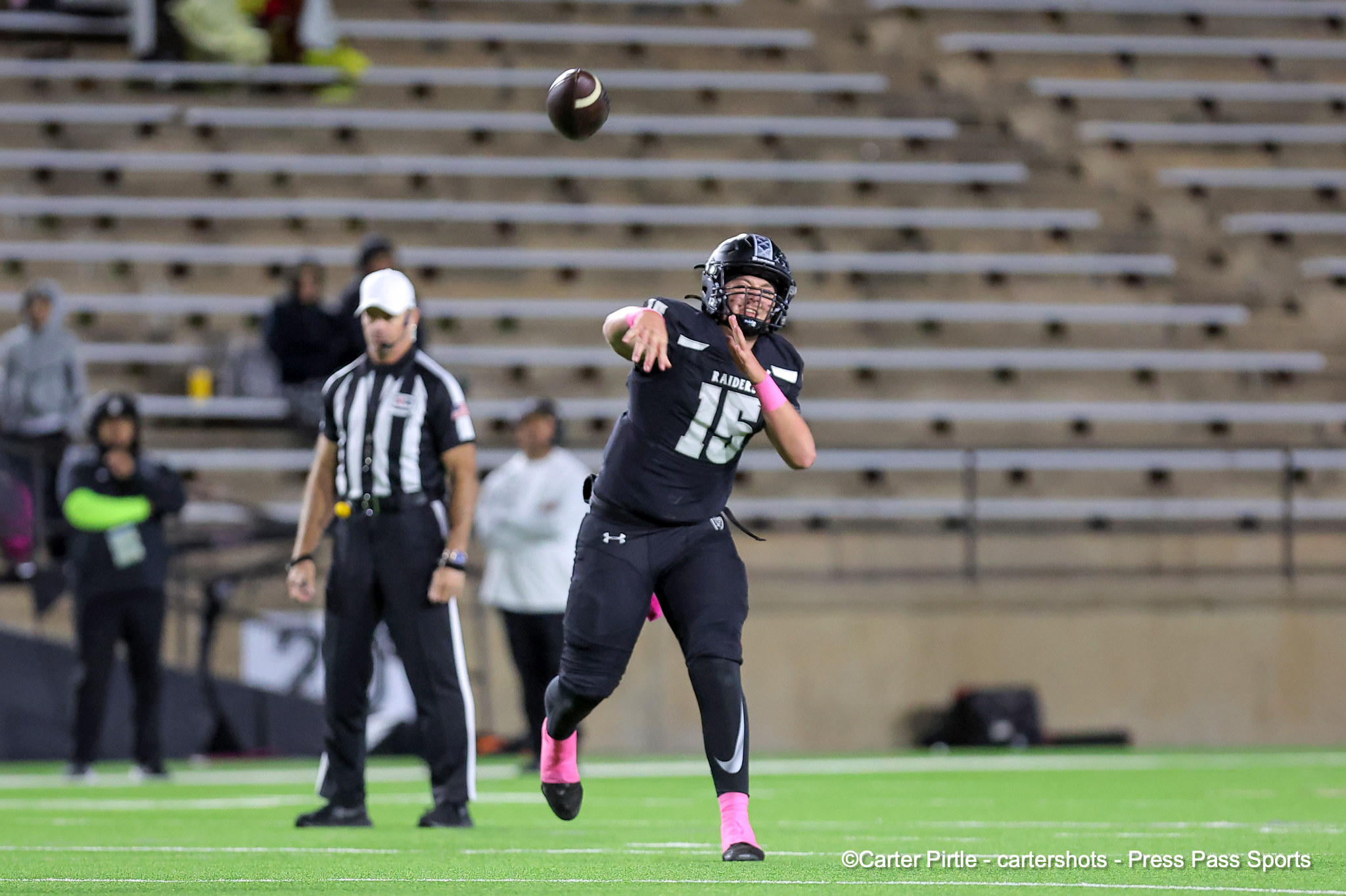 Football quarterback in black uniform throws a pass on a green indoor field, pink accessories, helmet on, with officials and spectators in the background.