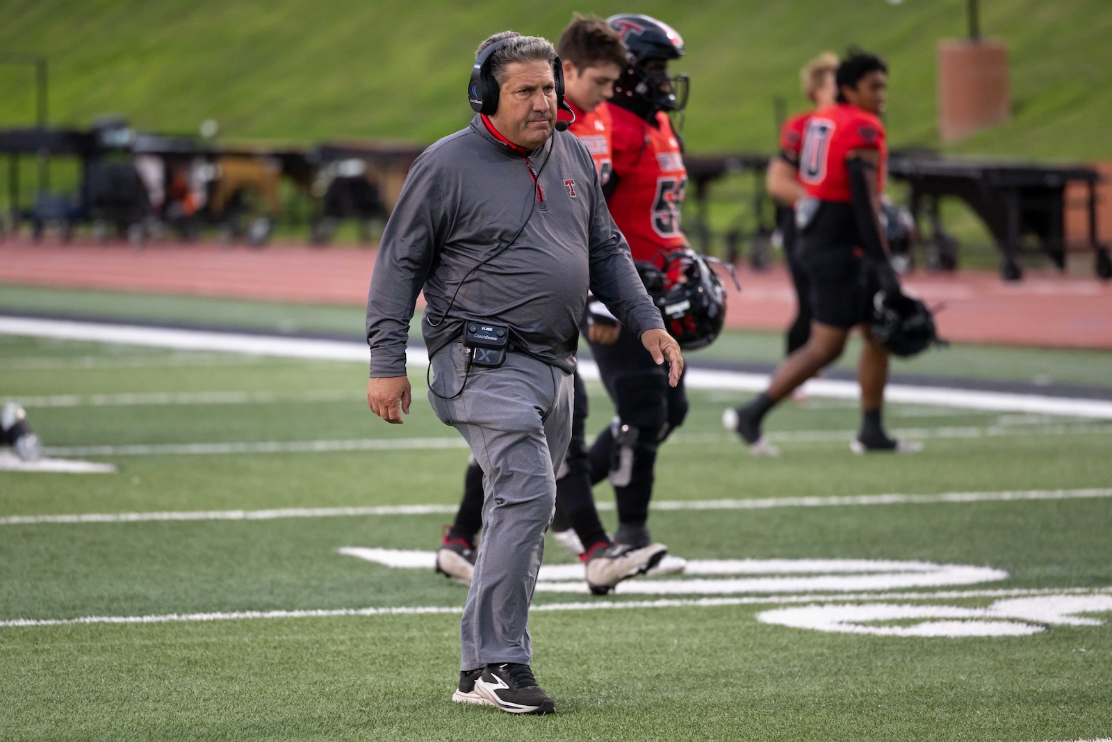 MHS-THS-FB-1 | Press Pass Sports Coach in a gray tracksuit with a headset walks across a football field, players in red jerseys blurred in the background.