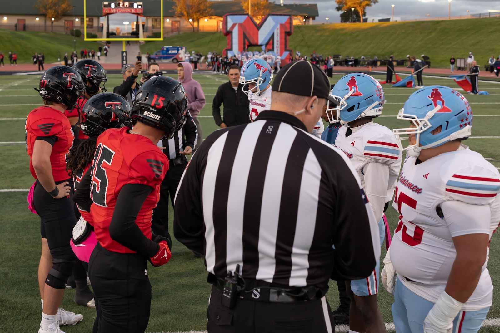 MHS-THS-FB-2 | Press Pass Sports Two football teams facing off at midfield with a referee in a black-and-white striped shirt ready to coin toss on a sunny field.