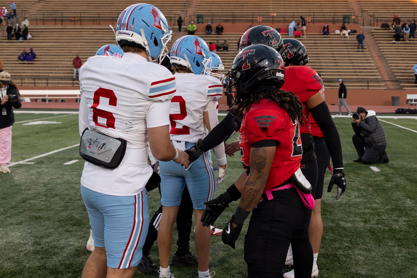 MHS-THS-FB-4 | Press Pass Sports Football players in white and light blue uniforms shake hands with players in red and black uniforms on a football field before a game, stands in the background.