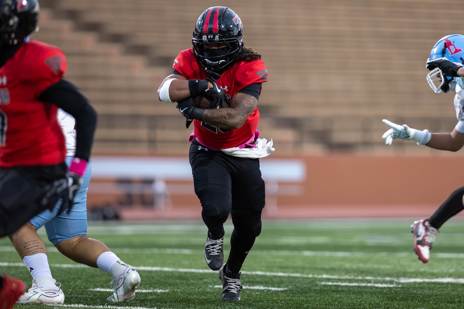MHS-THS-FB-5 | Press Pass Sports Football player in a red jersey runs with the ball tucked under his arm on a green field during a game.