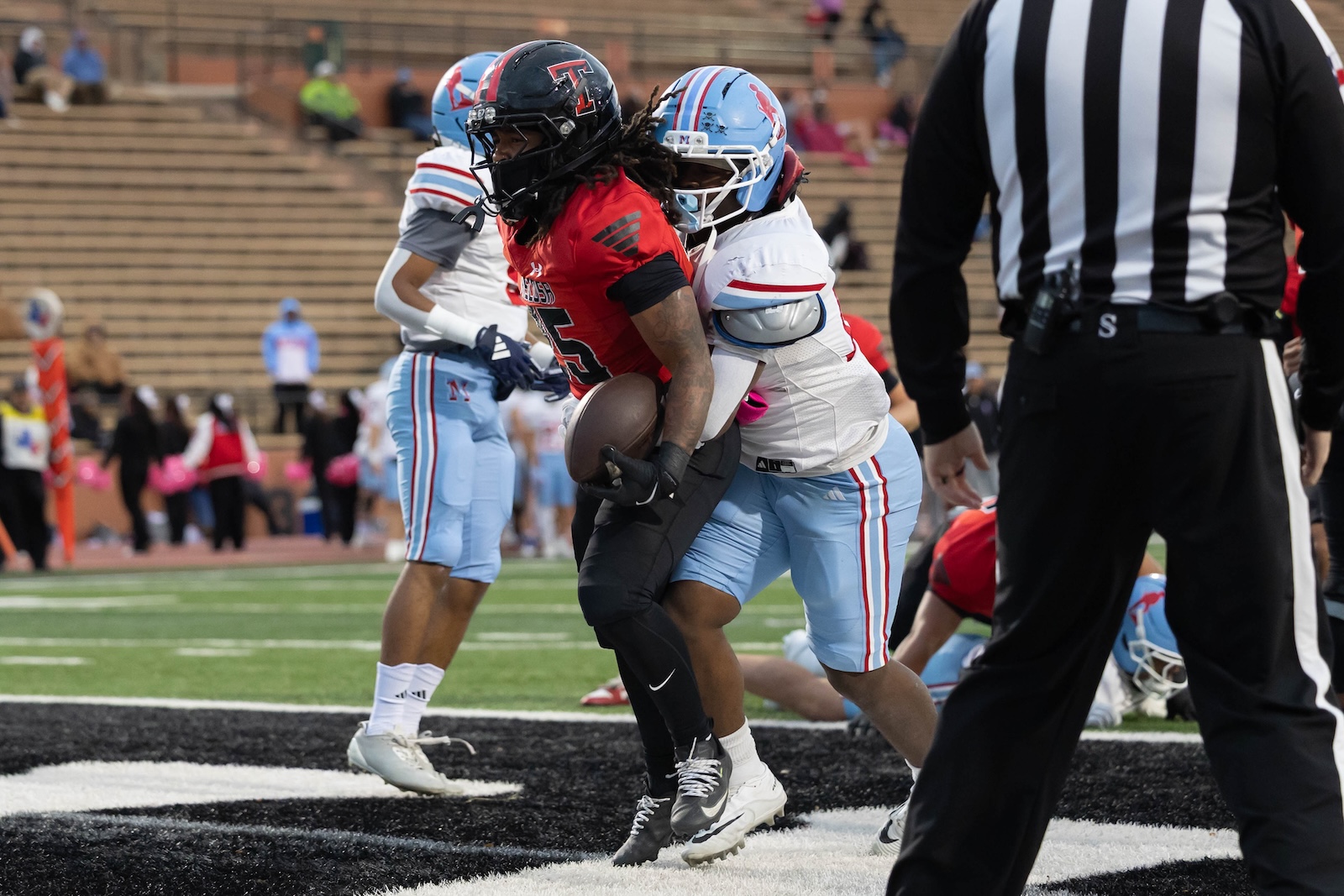 MHS-THS-FB-6 | Press Pass Sports Football play: a red-jersey runner holds the ball while a blue-jersey defender wraps his arms around him from behind near the end zone marker. Referee visible on the right.