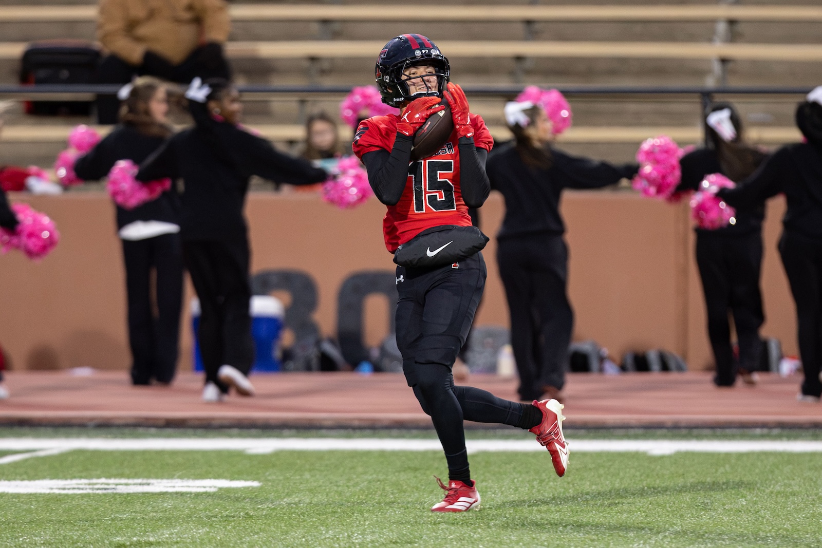 MHS-THS-FB-9 | Press Pass Sports Football player in red jersey #15 catching a ball on a turf field, with cheerleaders in black and pink pom-poms in the background