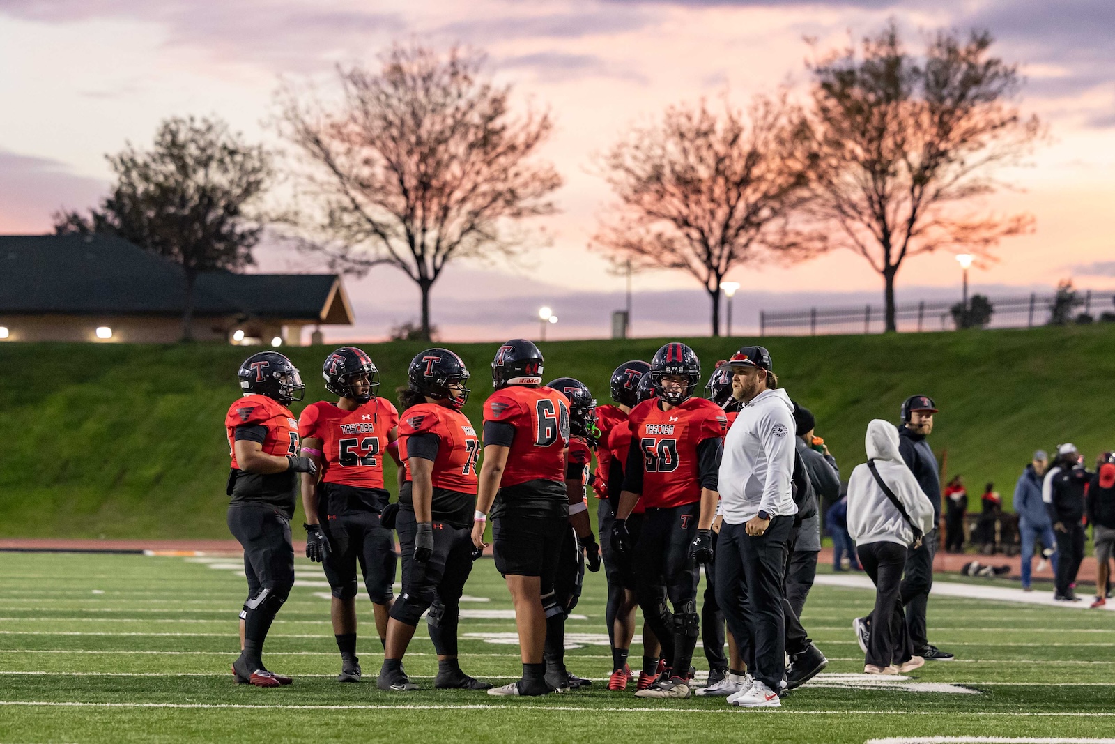 MHS-THS-FB-10 | Press Pass Sports Football team in red jerseys huddles on a green field at sunset; coach in white hoodie nearby.