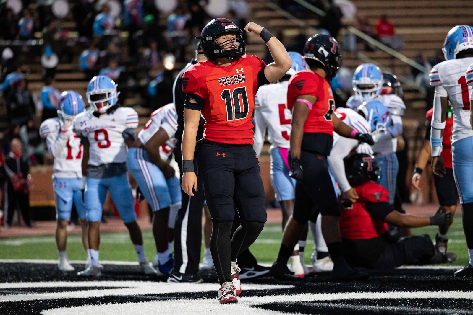 MHS-THS-FB-11 | Press Pass Sports Football player in red jersey with number 10 flexing his arm on a football field, surrounded by teammates and opponents.