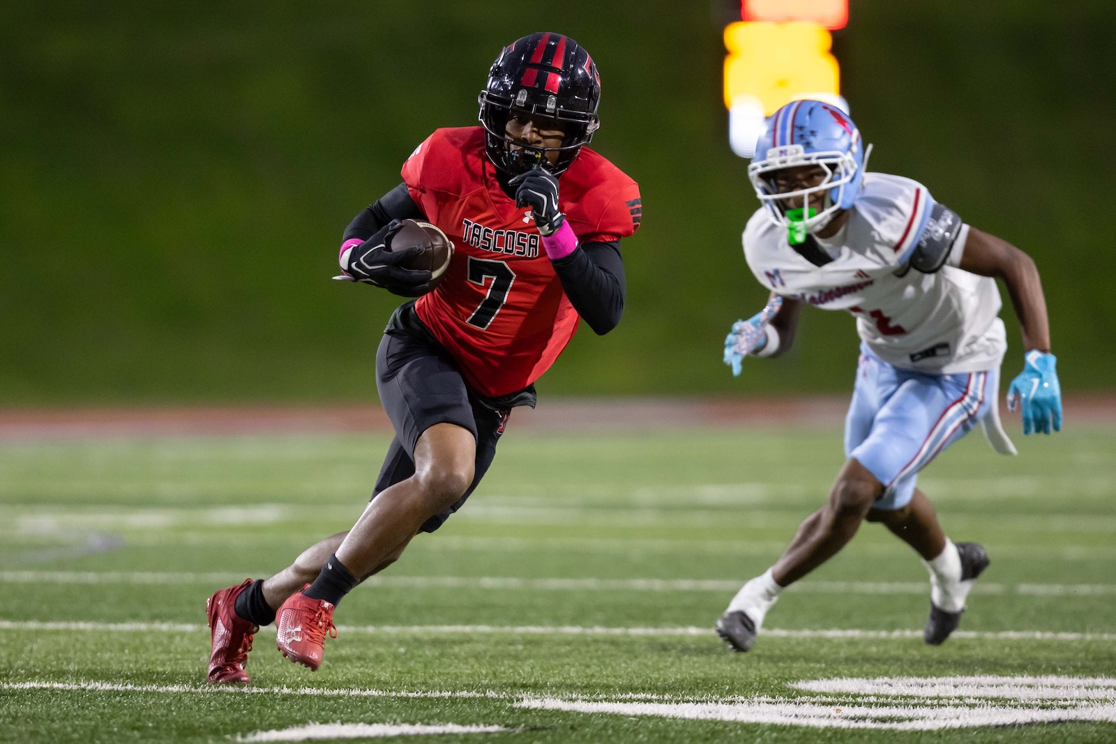 MHS-THS-FB-13 | Press Pass Sports A football player in a red Tasosa jersey with number 7 runs with the ball, pursued by a defender in white and blue under stadium lights.