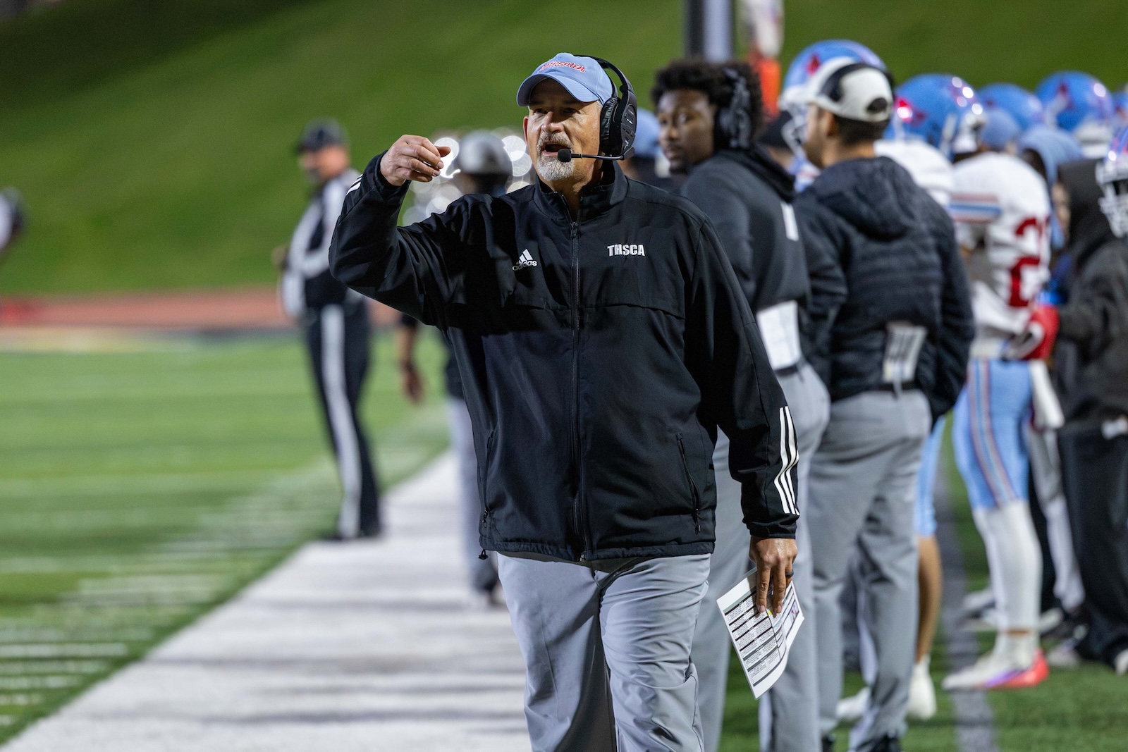 MHS-THS-FB-19 | Press Pass Sports Football coach with headset gesturing on the sideline during a game, wearing a black jacket and gray pants, holding a play sheet.