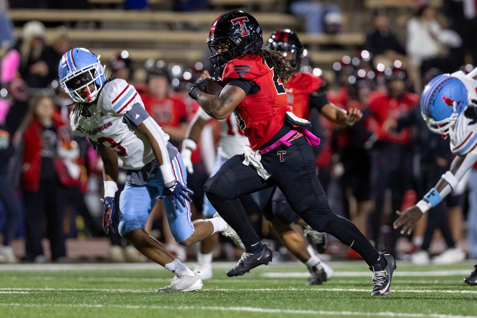 MHS-THS-FB-20 | Press Pass Sports A football player in red and black uniform runs with the ball, chased by a defender in white and blue on a green turf field.