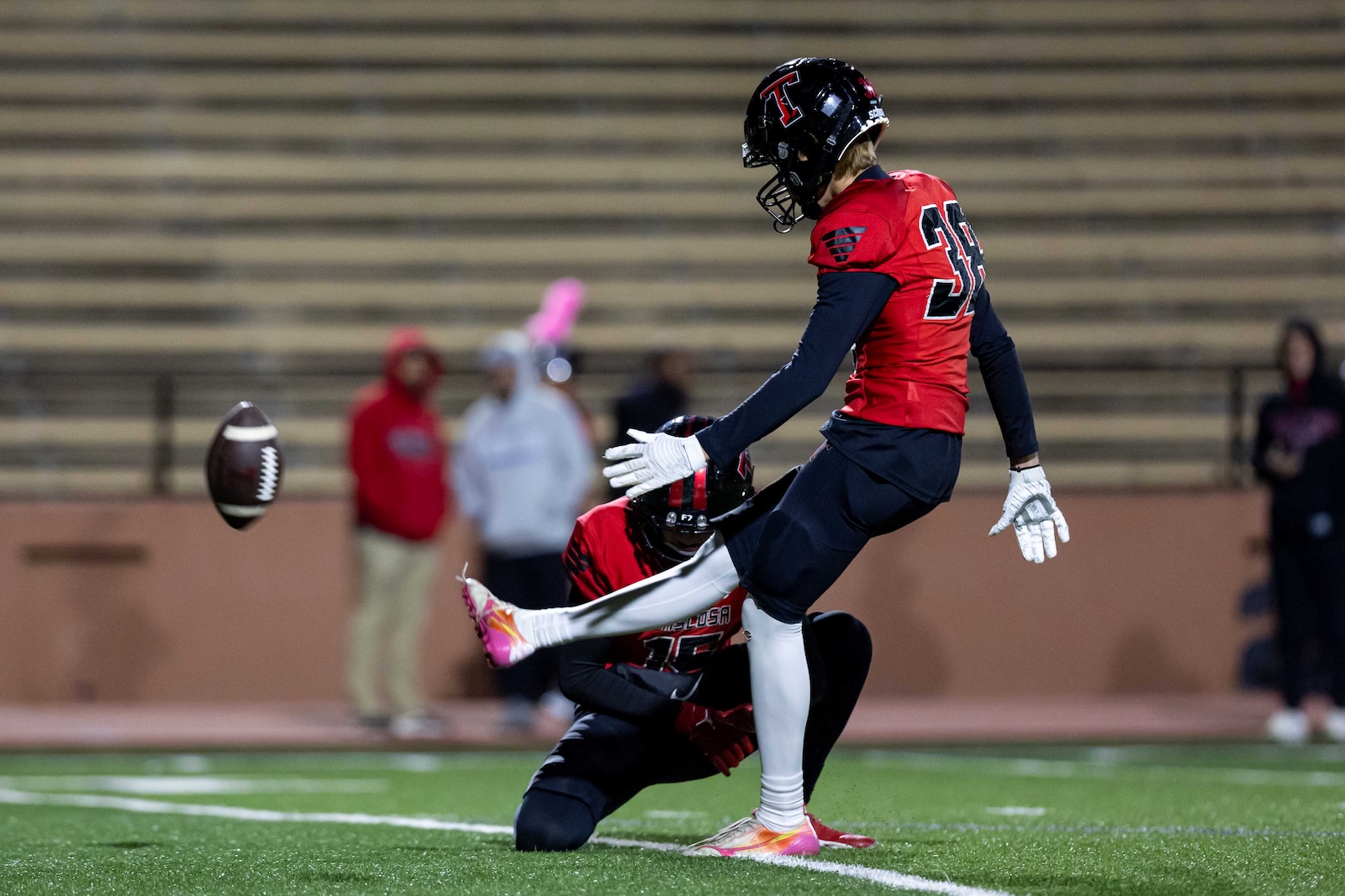 MHS-THS-FB-22 | Press Pass Sports Football player in a red jersey leaps to kick the football during a night game on a green turf field.