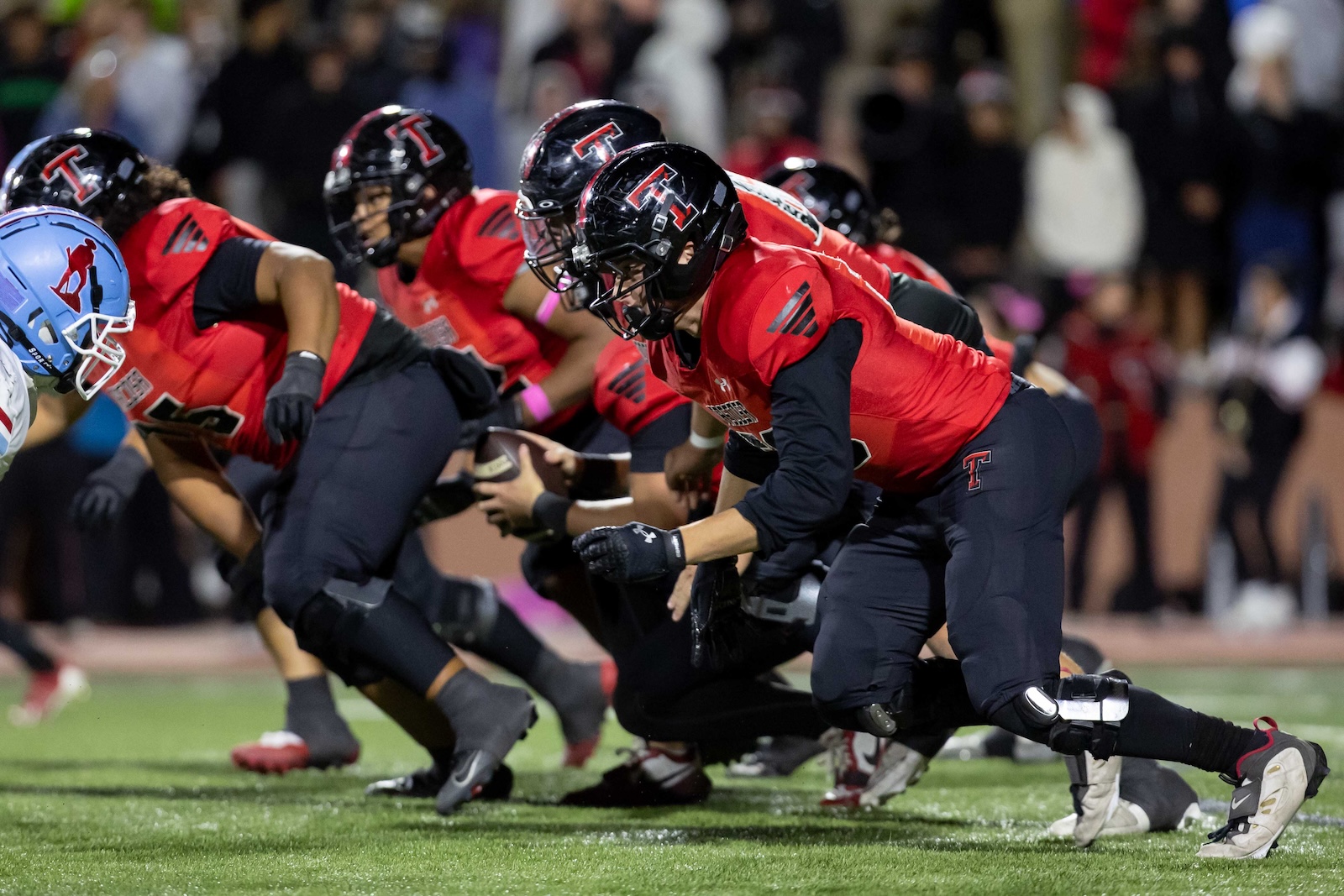 MHS-THS-FB-25 | Press Pass Sports American football players in red jerseys crouch at the line of scrimmage as a play begins, with a crowd in the stands behind them.