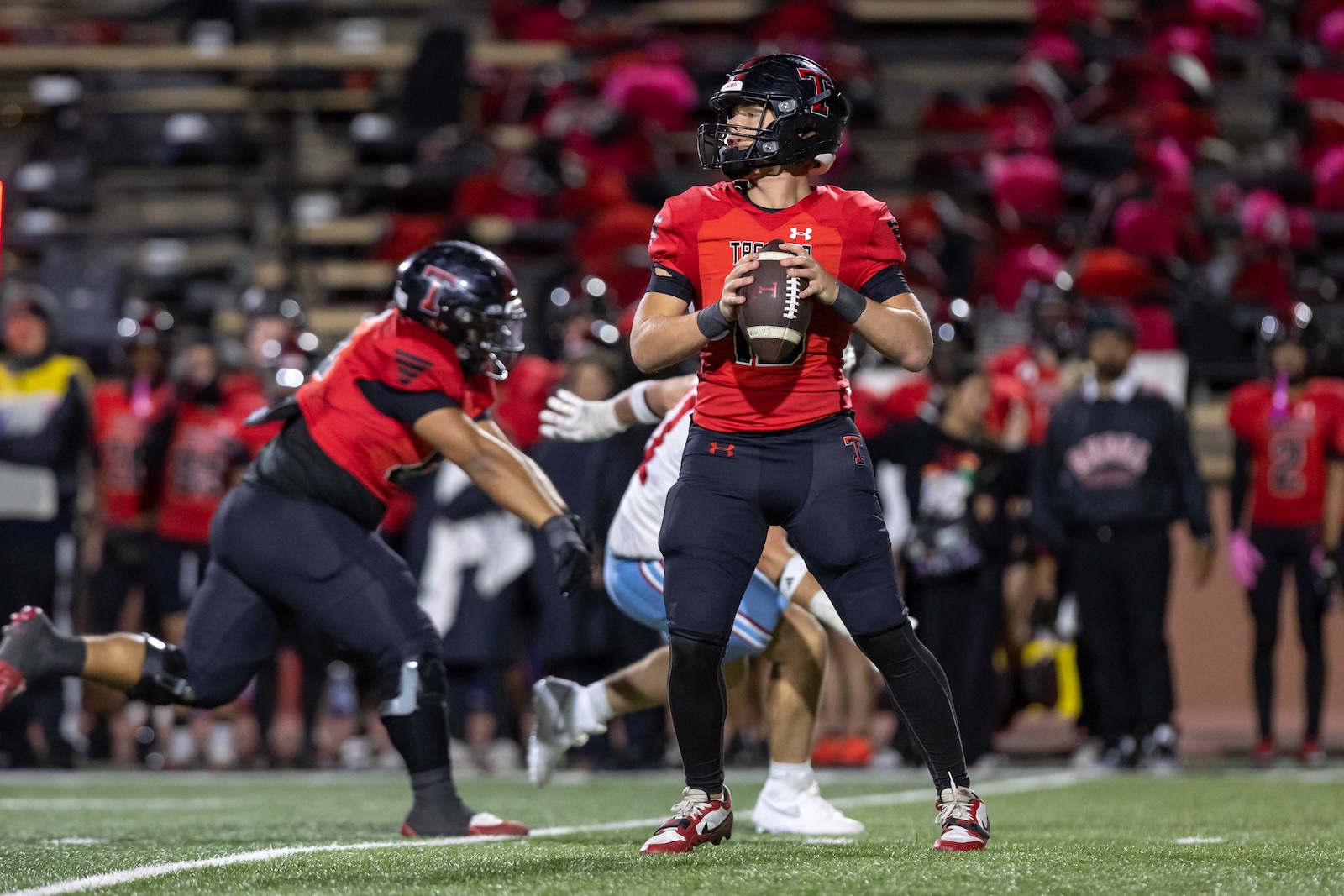 MHS-THS-FB-28 | Press Pass Sports Quarterback in red jersey poised to throw a football during a game, with defenders and spectators in the background.