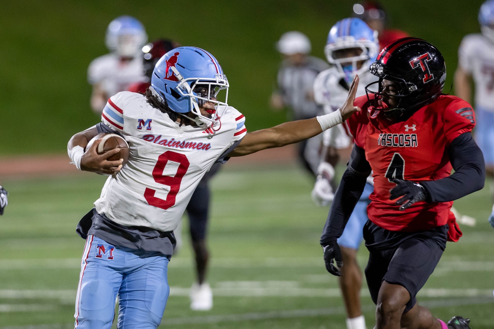 MHS-THS-FB-30 | Press Pass Sports Football player in white and light blue with the ball, number 9, extended arm reaching toward a defender in red number 4 on a green field stadium.”,