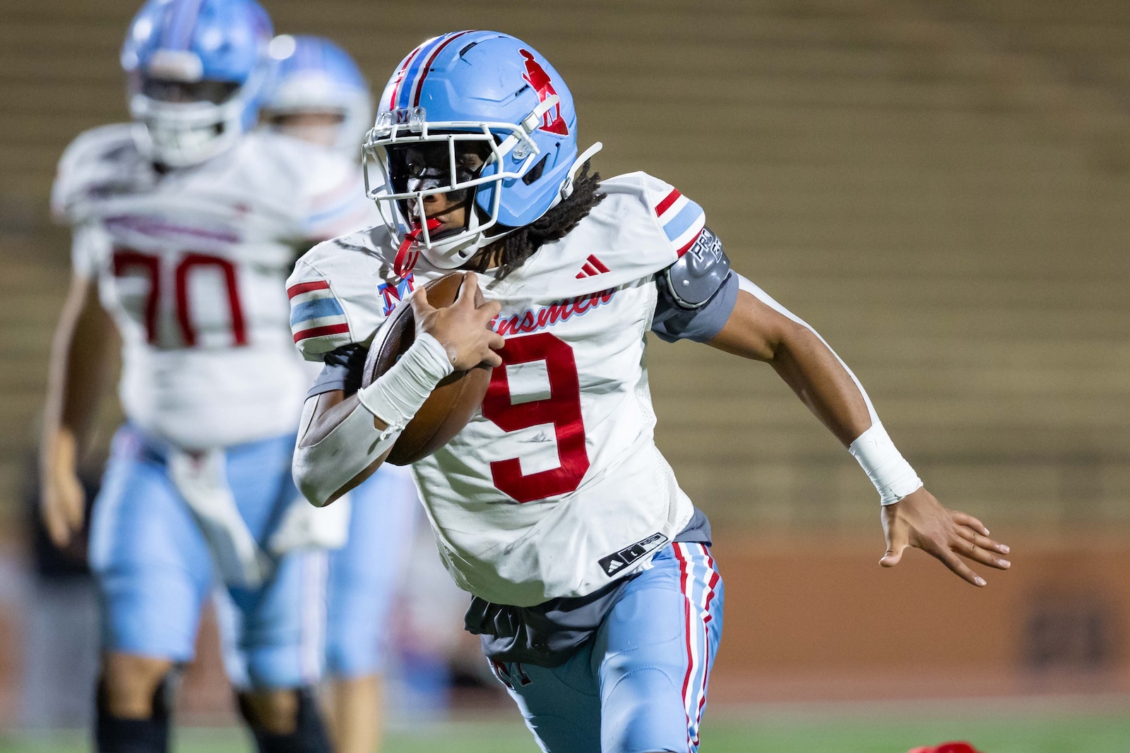 MHS-THS-FB-31 | Press Pass Sports Football player wearing a white jersey with number 9 and a blue helmet runs with the ball during a game, with teammates in the background.