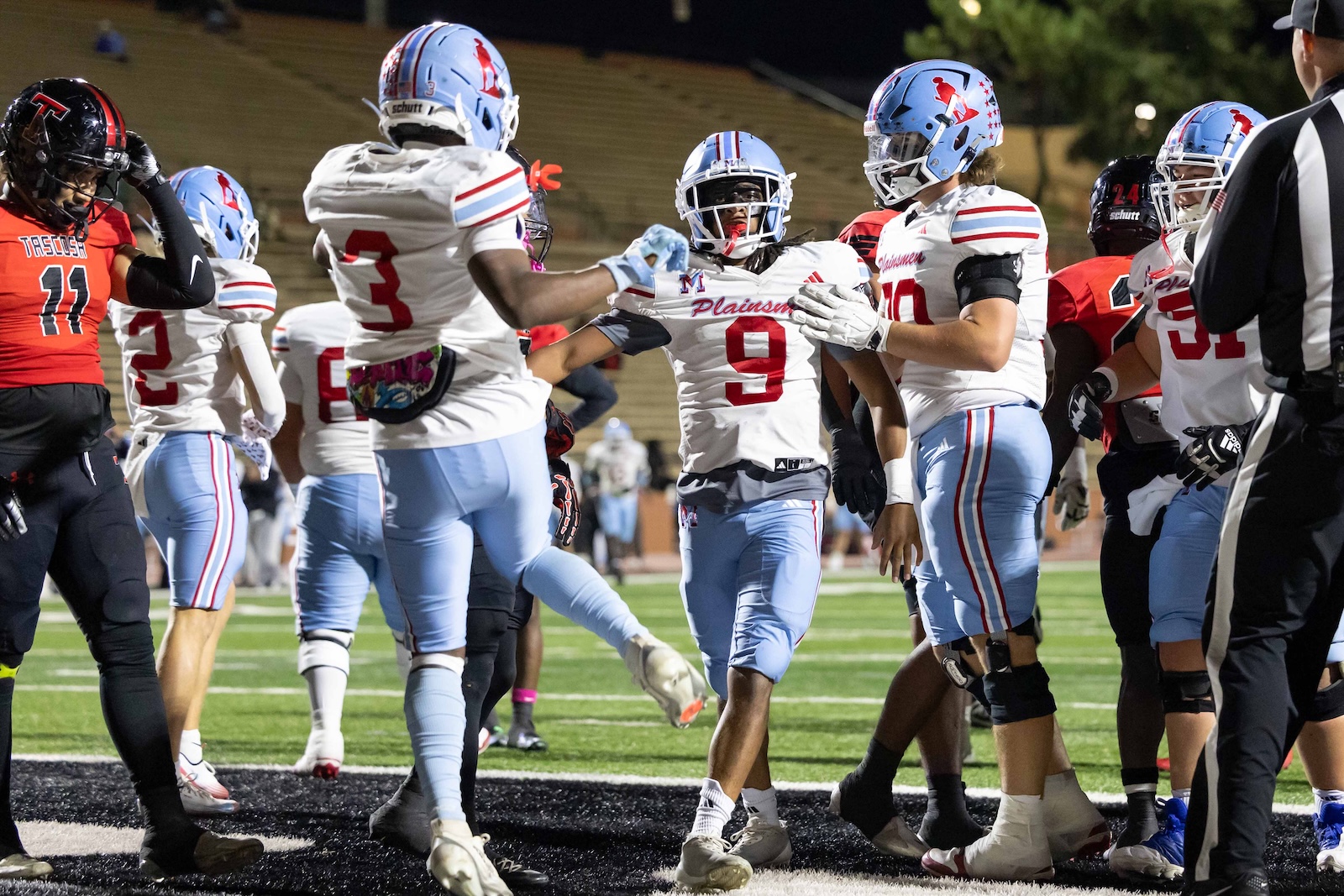MHS-THS-FB-32 | Press Pass Sports Football players in white and light‑blue Plainsmen uniforms high‑five on a turf field at night after a play attack zone touch.