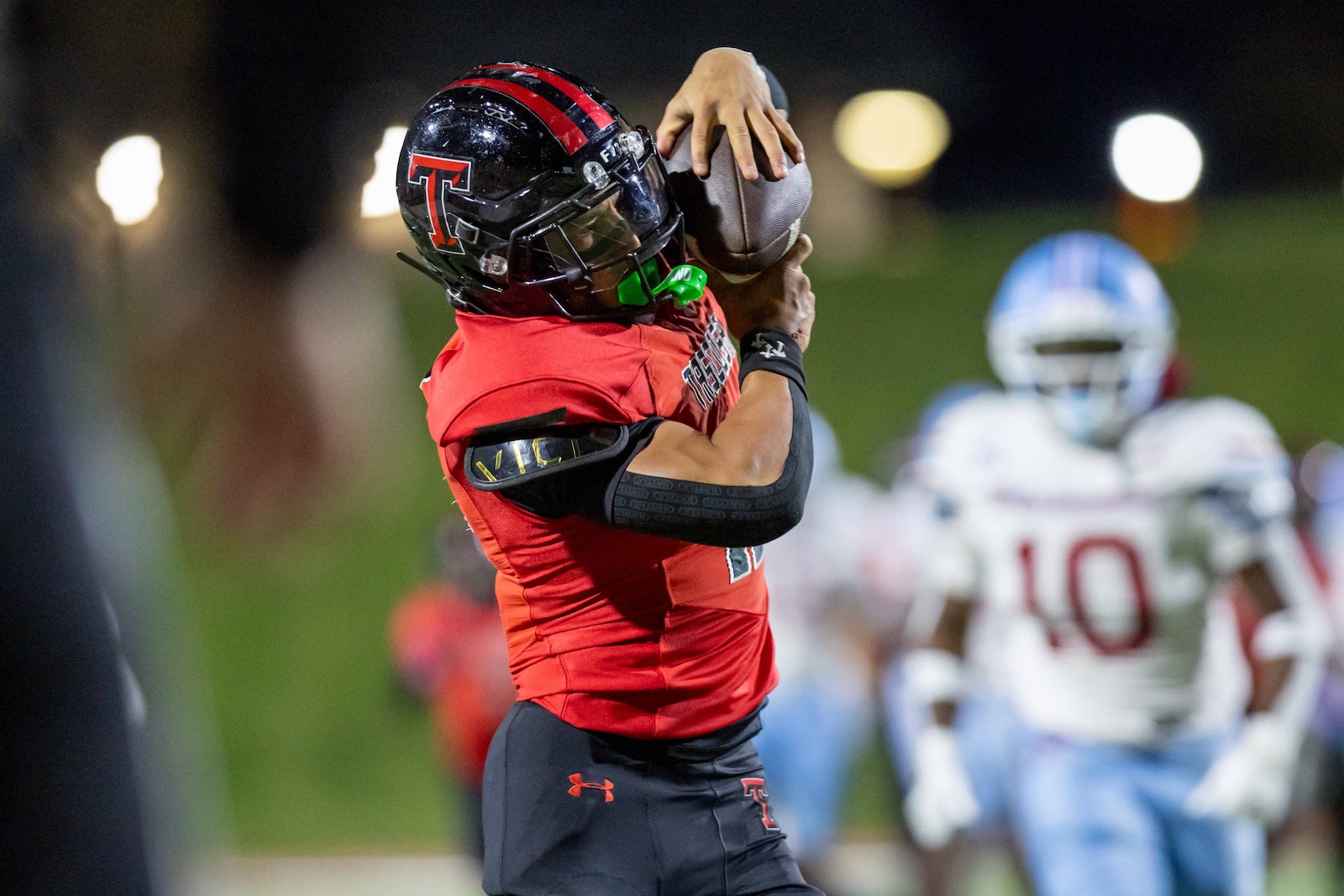 MHS-THS-FB-34 | Press Pass Sports Football player in red jersey leaps to catch a pass, with defenders blurred in the background under stadium lights.
