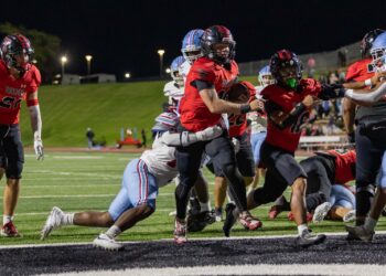 MHS-THS-FB-39 | Press Pass Sports Running back in a red jersey salvages the ball and charges forward as defenders close in during a night football game on a lit field.