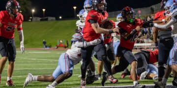 Running back in a red jersey salvages the ball and charges forward as defenders close in during a night football game on a lit field.