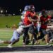 MHS-THS-FB-39 | Press Pass Sports Running back in a red jersey salvages the ball and charges forward as defenders close in during a night football game on a lit field.