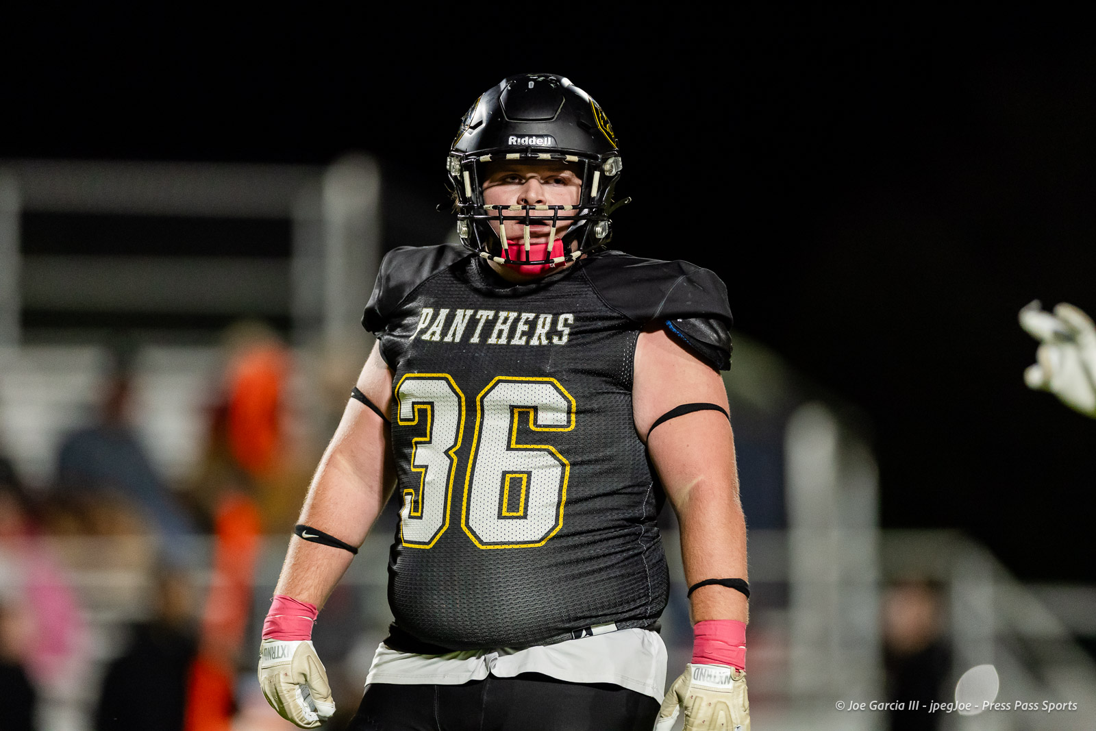 groom-vs-follett-football-jpegJoe-pps-12 | Press Pass Sports Football player in a black Panthers jersey #36, wearing a helmet and pink gloves, on a night football field.