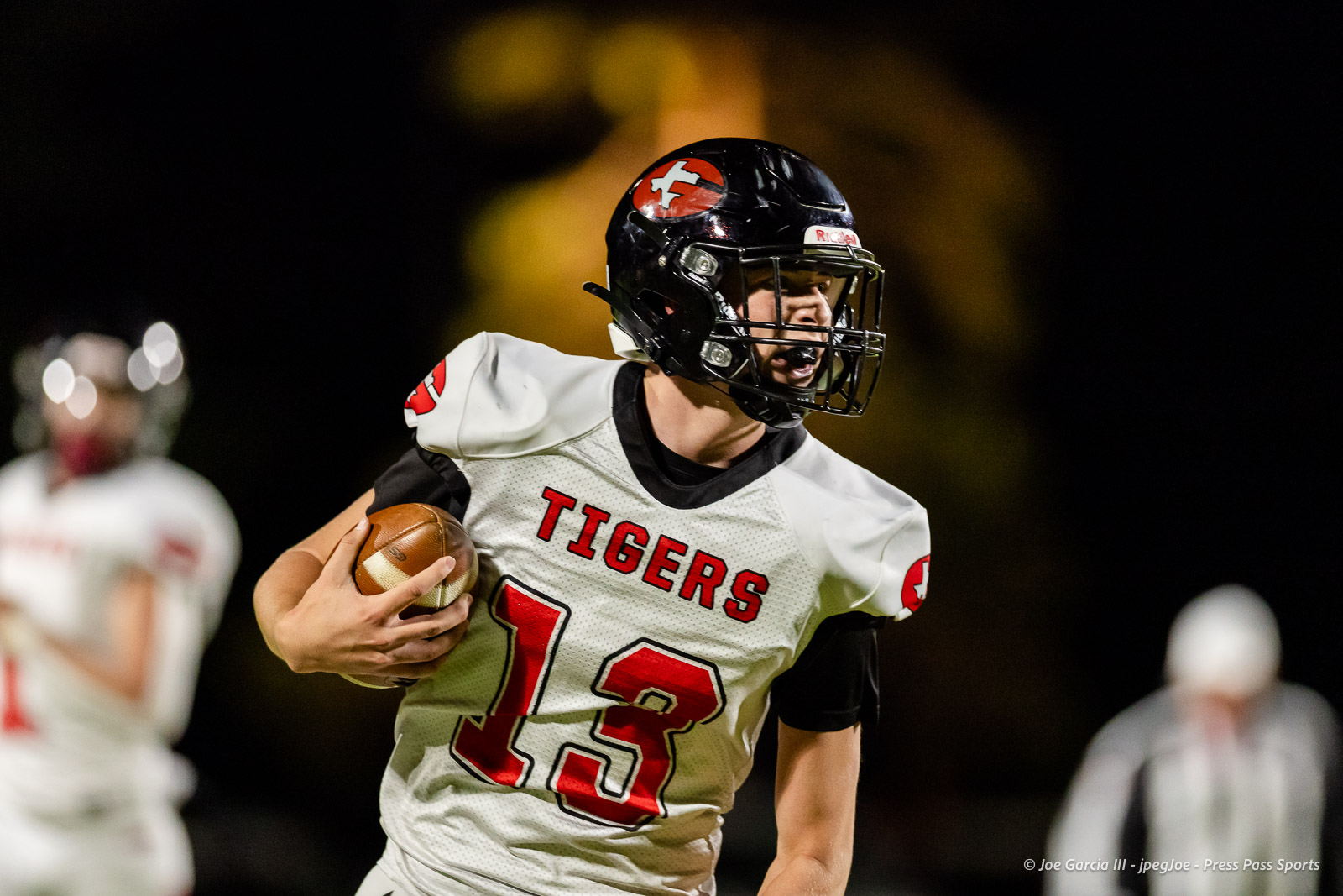 groom-vs-follett-football-jpegJoe-pps-15 | Press Pass Sports Football player in white Tigers jersey (#13) runs with the ball during a night game, helmet on, focused ahead.