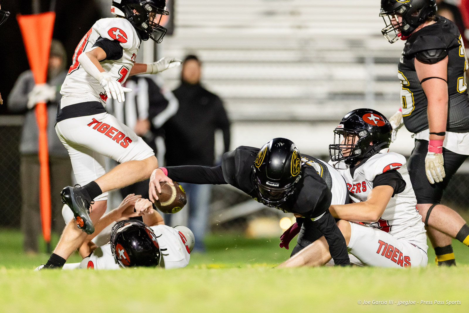 groom-vs-follett-football-jpegJoe-pps-19 | Press Pass Sports Football players in white Tigers uniforms tackle a black-uniform opponent on a grassy field; a loose football lies nearby.