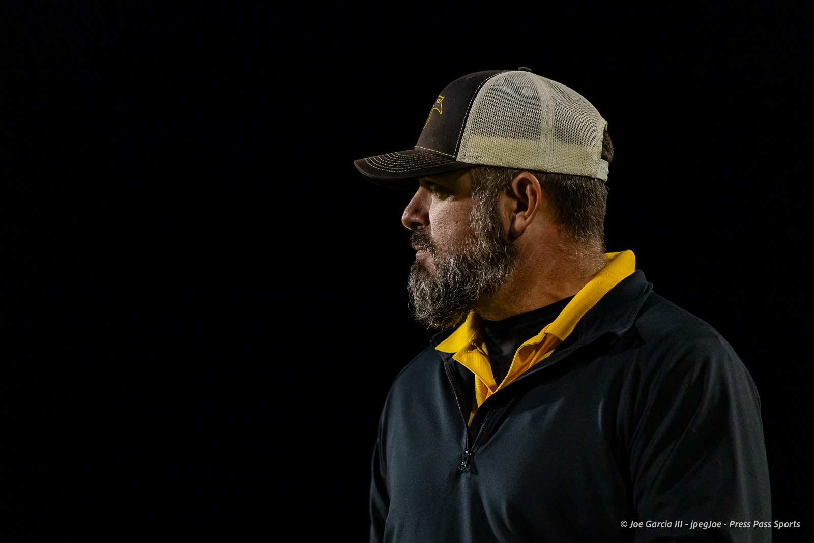 groom-vs-follett-football-jpegJoe-pps-20 | Press Pass Sports Profile of a man with a gray beard wearing a black cap and a yellow-collared polo under a dark jacket against a black background, looking to the left with a neutral expression.