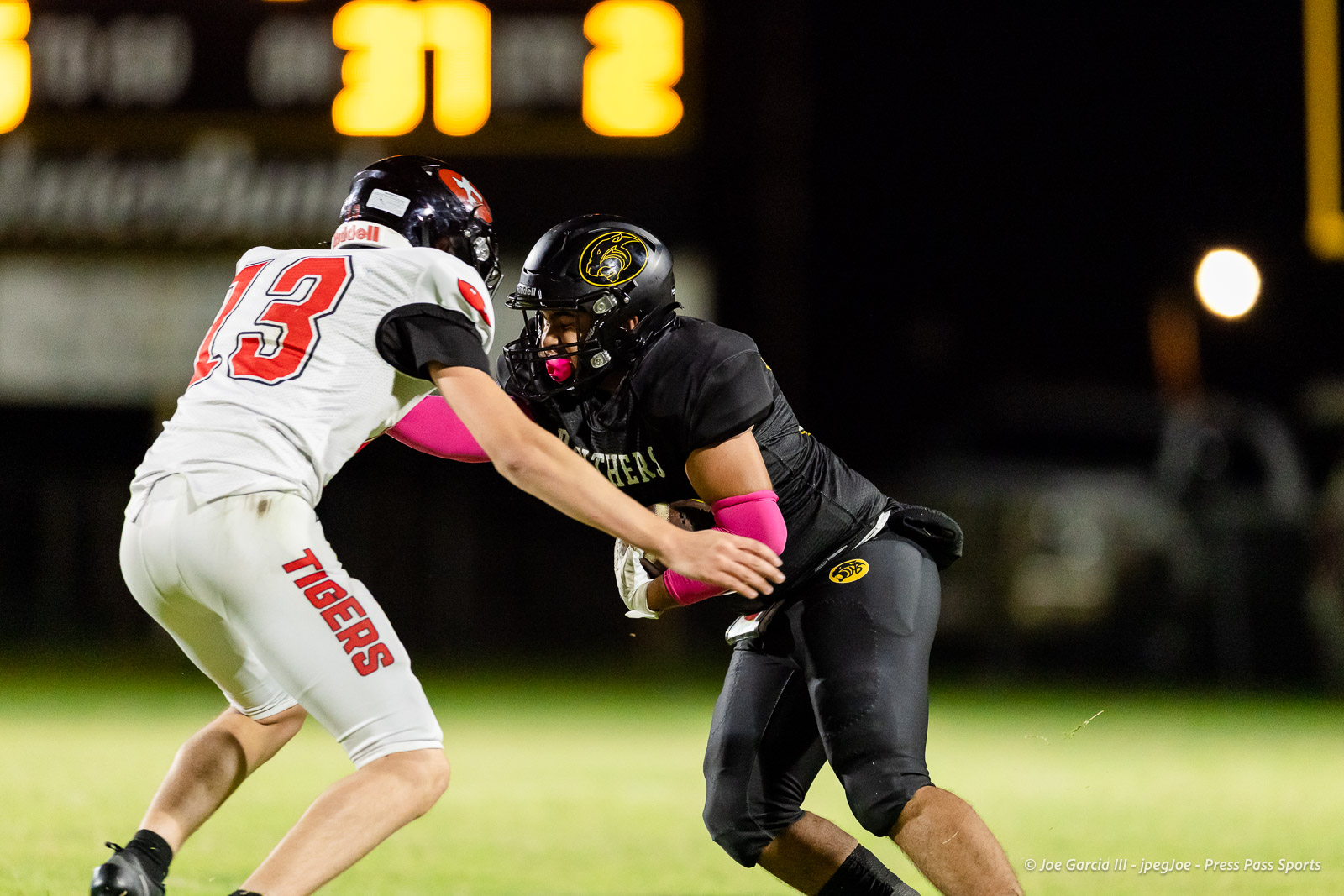 groom-vs-follett-football-jpegJoe-pps-21 | Press Pass Sports Two football players collide at night; white Tigers jersey (number 23) vs black-uniformed runner with pink accessories under stadium lights.