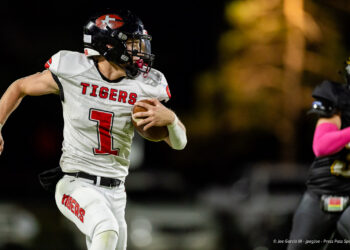 groom-vs-follett-football-jpegJoe-pps-23 | Press Pass Sports Football player in white 'Tigers' jersey runs with ball during night game, aimed forward with helmet and red number 1 visible on chest.