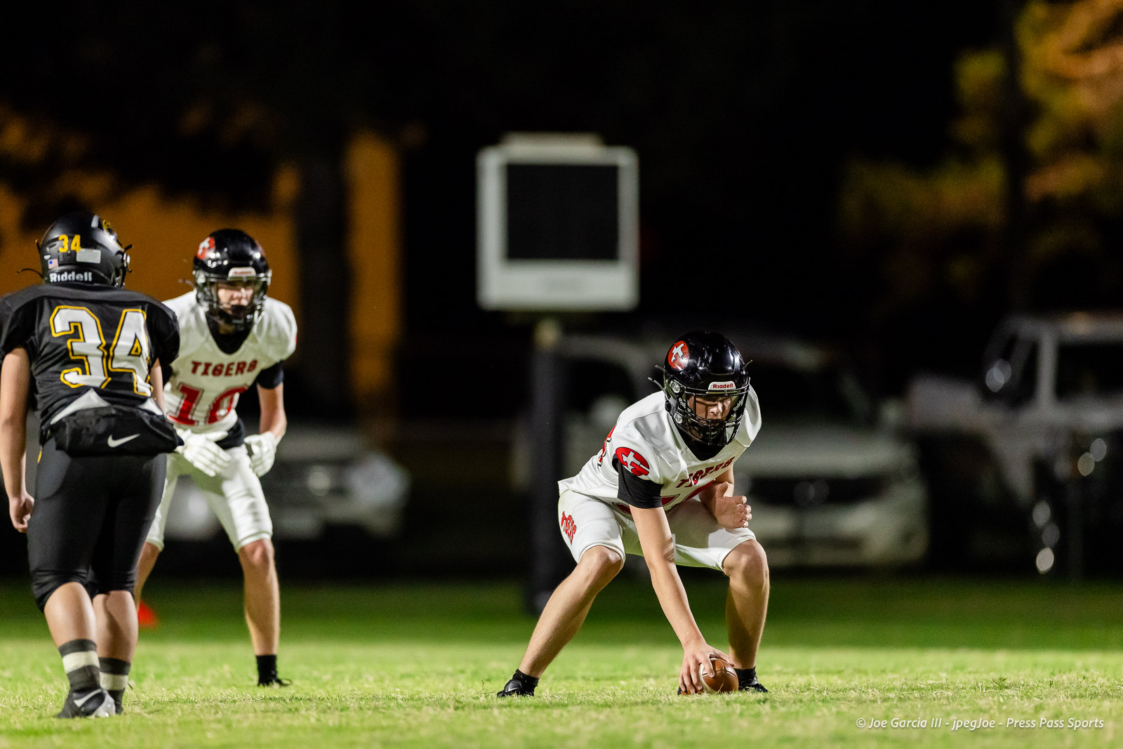 groom-vs-follett-football-jpegJoe-pps-24 | Press Pass Sports Crouched white-team player with'TIGERS' on jersey and ball at the snap on a night football field, with defenders in black nearby and a blurred scoreboard in the background.