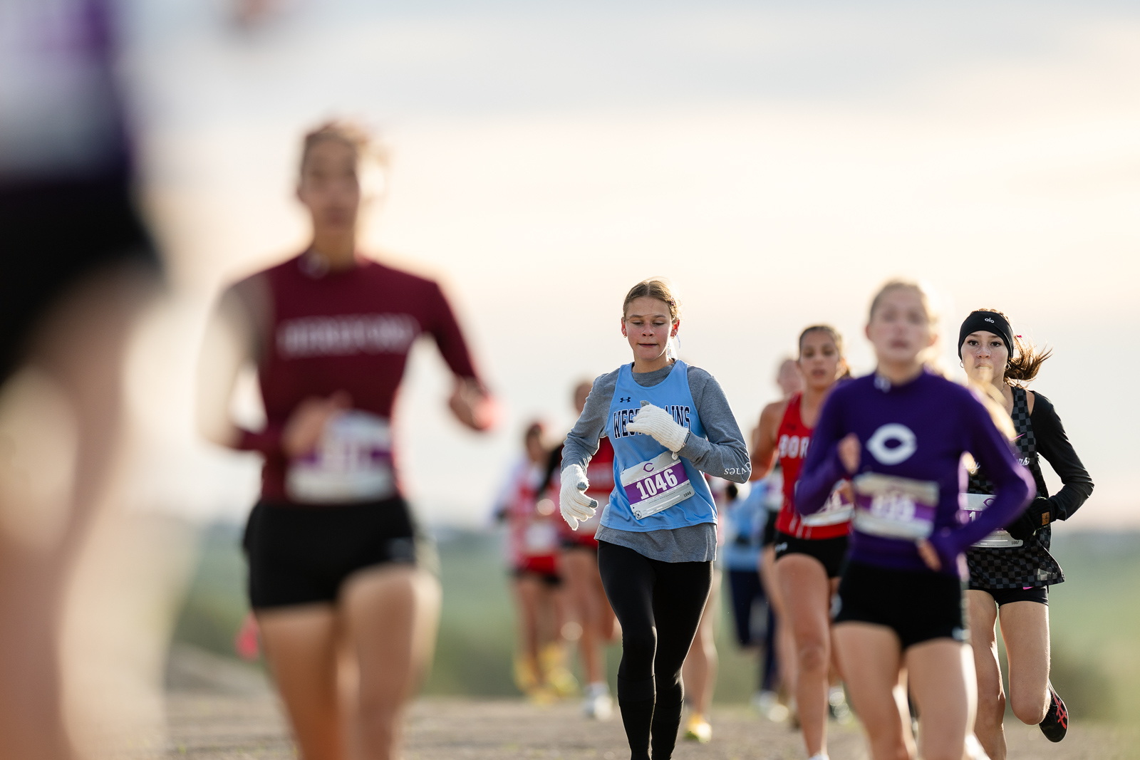 3A-4A-district-xc-jpegJoe-pps-5 | Press Pass Sports A young runner in a light blue top with bib 1046 leads a group of joggers on a sunny outdoor course, others blurred in the background.