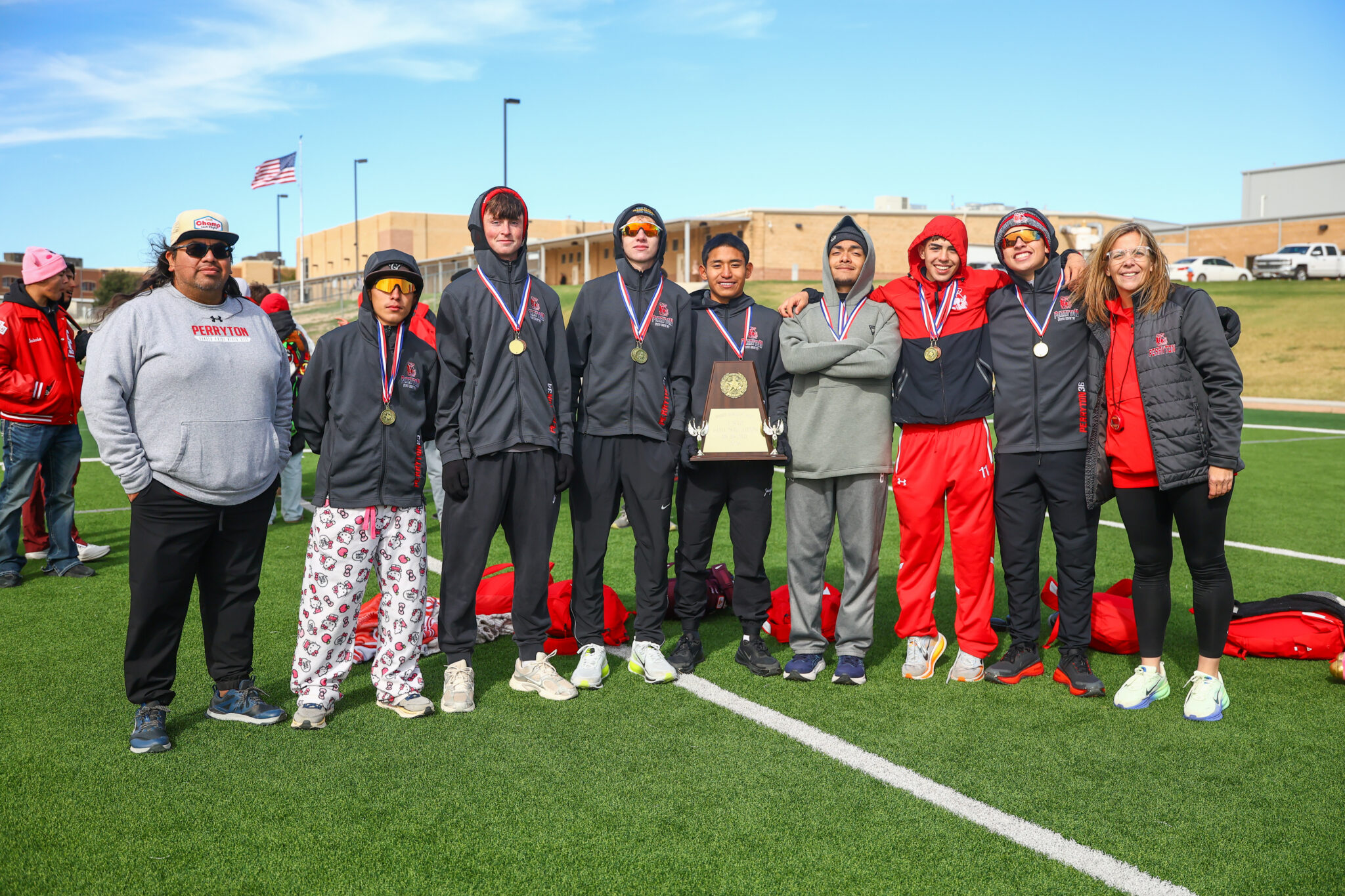 CT9A3377 | Press Pass Sports Group of smiling athletes with medals and a trophy on a sports field, wearing matching dark jackets.