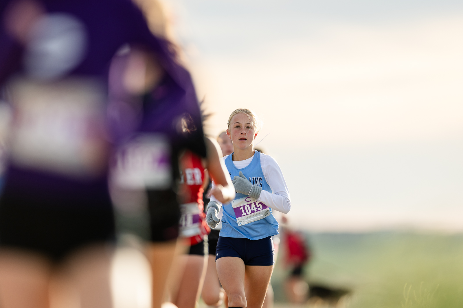 3A-4A-district-xc-jpegJoe-pps-7 | Press Pass Sports Female runner in a light blue top and navy shorts with bib 1045 competes in a race, others blurred in the foreground on a sunny day.