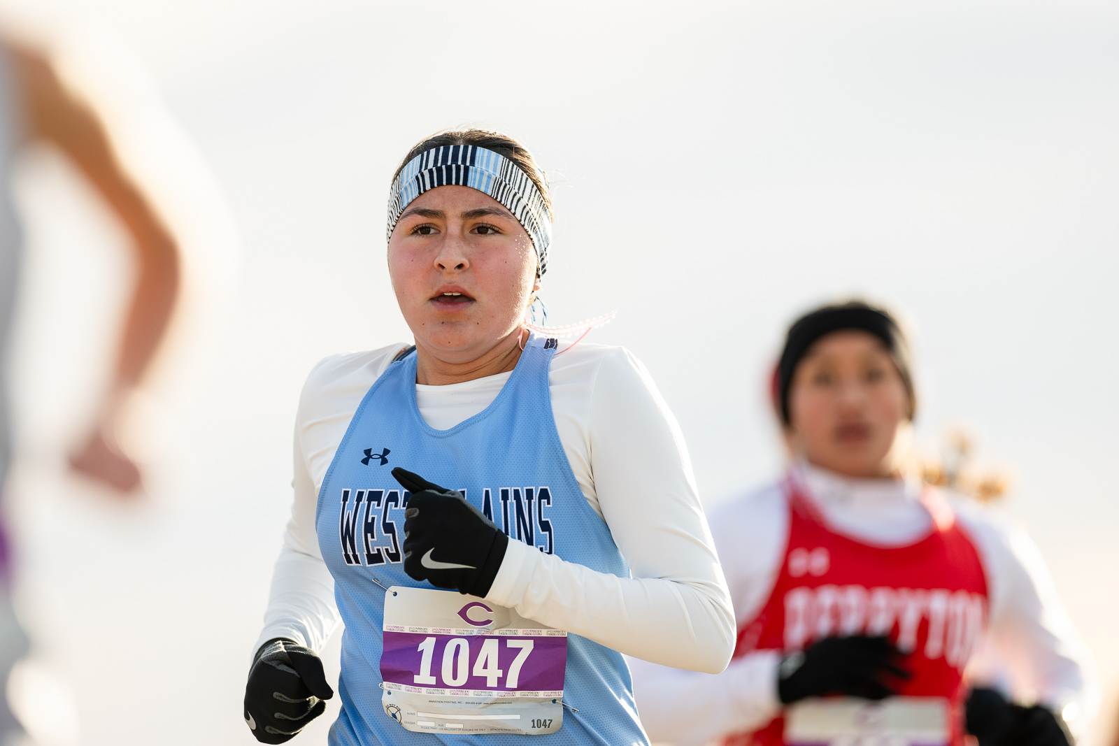 3A-4A-district-xc-jpegJoe-pps-8 | Press Pass Sports Female runner in a light blue jersey with bib 1047, striped headband and black gloves, in a race with a blurred red-jersey competitor behind her.