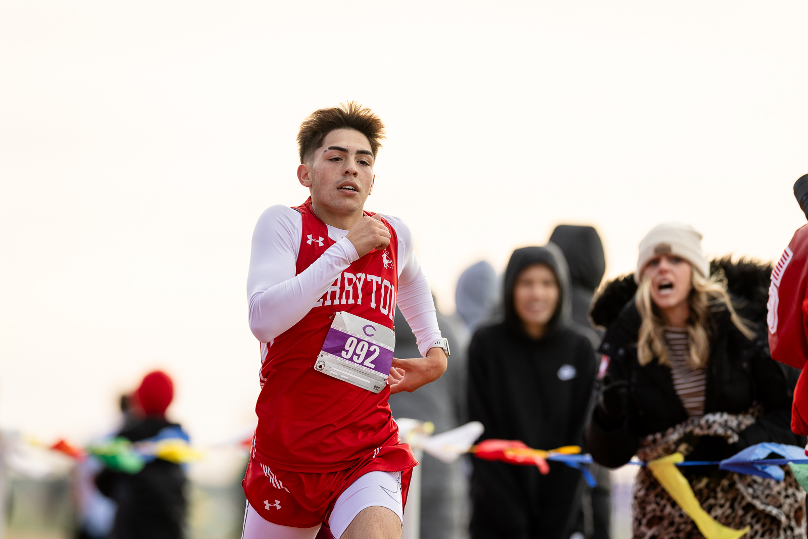 district-3a-4a-cross-country-jpegJoe-pps-92 | Press Pass Sports Male runner in a red uniform crossing the finish line, bib number 992, spectators cheering in the background.