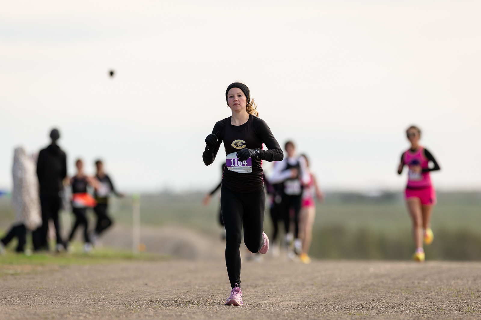 district-3a-4a-cross-country-jpegJoe-pps-133 | Press Pass Sports Female runner in black athletic gear and headband, bib 1104, sprinting on a gravel path with blurred runners in the background.