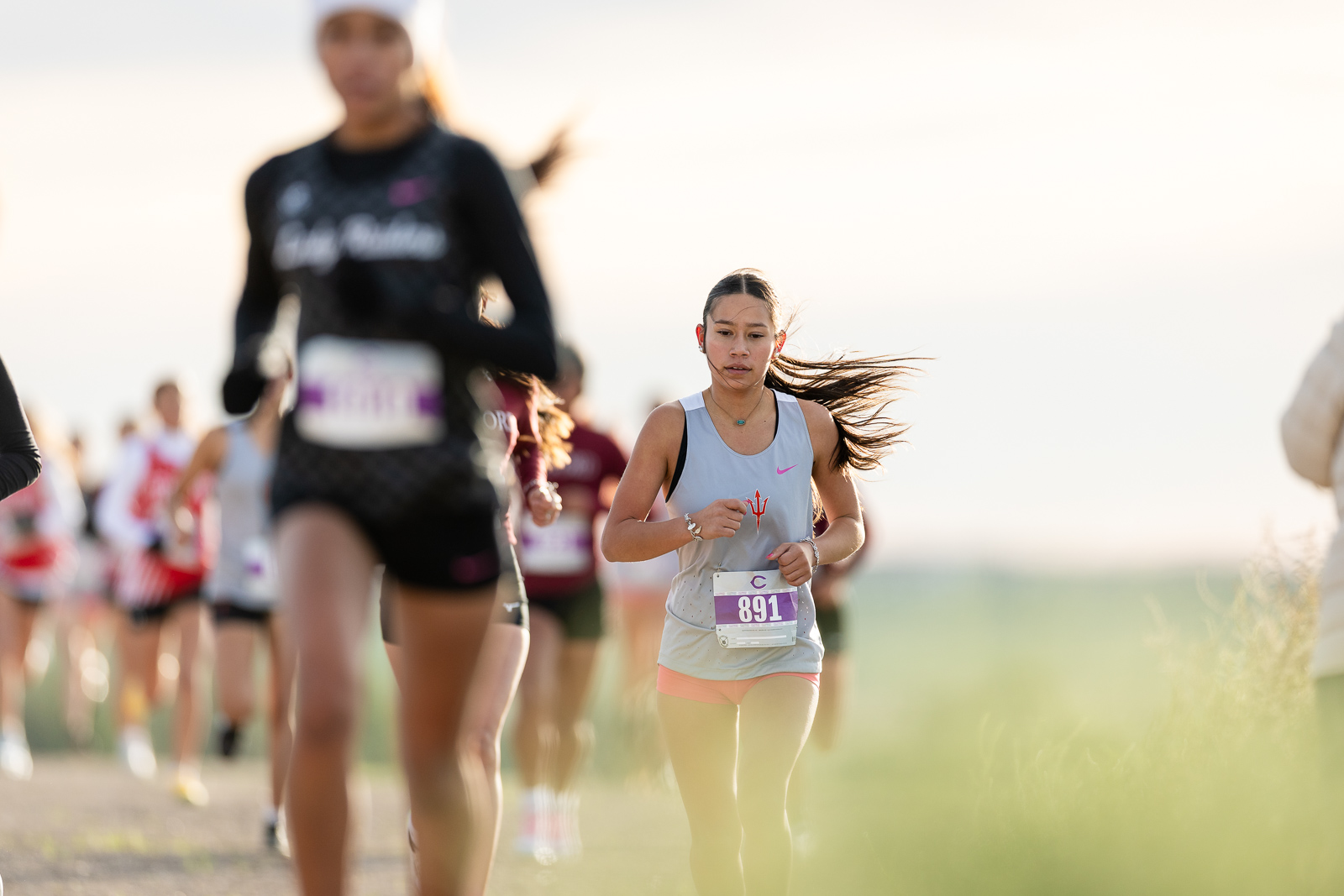 3A-4A-district-xc-jpegJoe-pps-10 | Press Pass Sports Female runner in a light gray tank top with bib 891 leads a group in a sunny race on a dirt path, foreground grasses blurred.