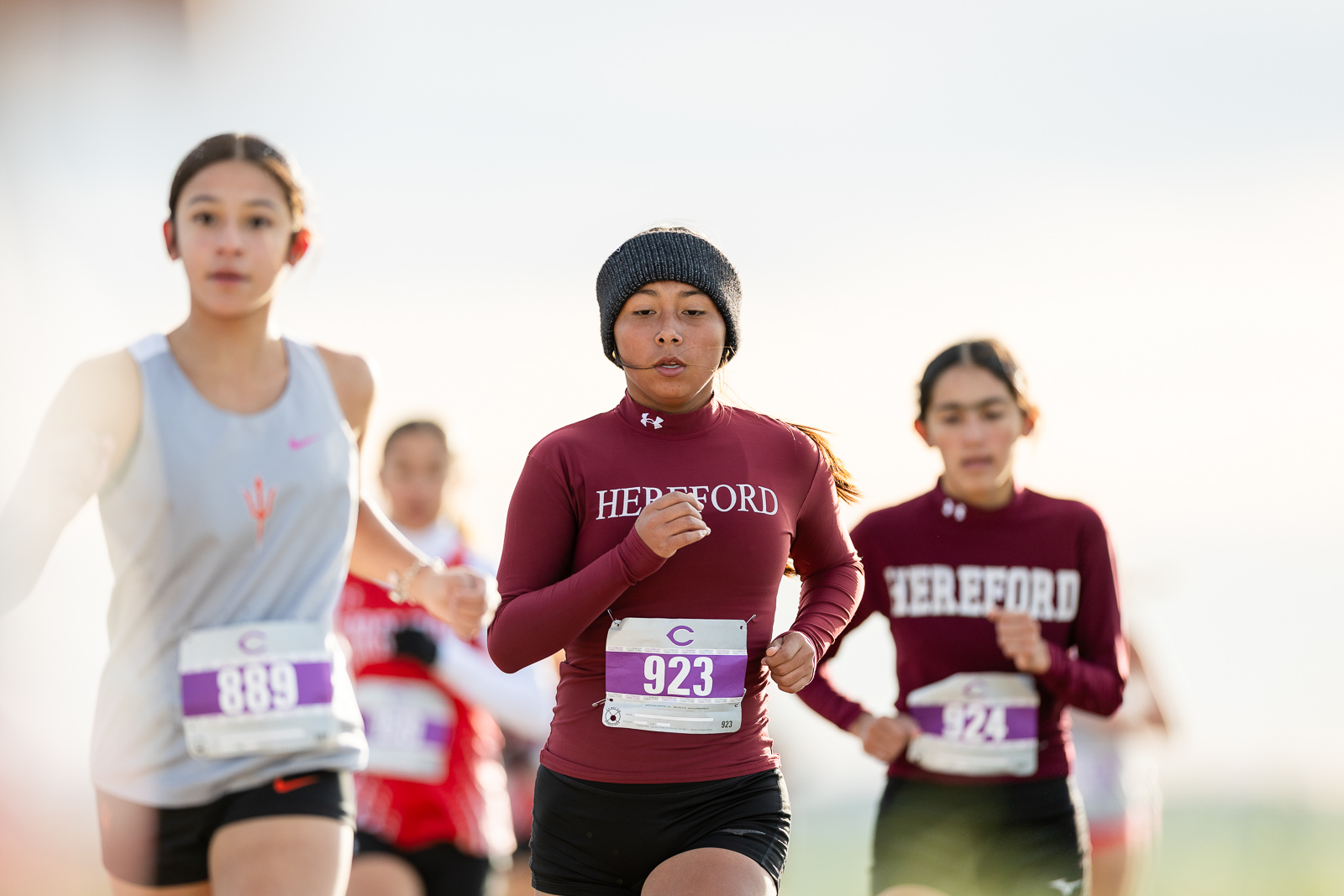 3A-4A-district-xc-jpegJoe-pps-11 | Press Pass Sports Female runners in a race, central athlete wearing a maroon Hereford top and knit cap, bib 923, running toward camera.